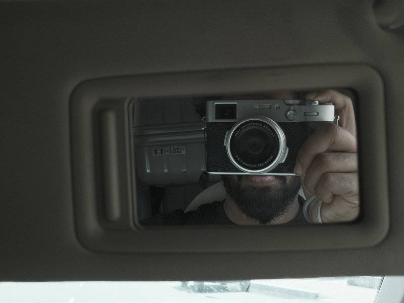 A person taking a selfie in a car's rearview mirror, showing a camera and part of their face.