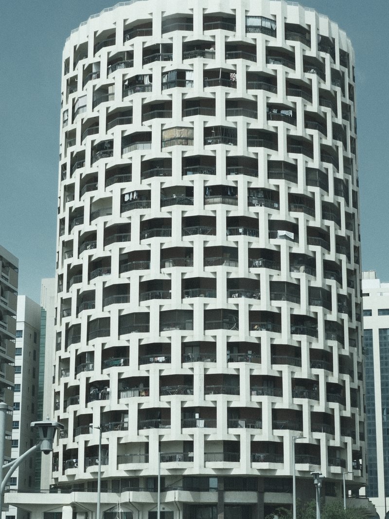 A modern cylindrical building with a unique lattice design, surrounded by other structures under a clear sky.