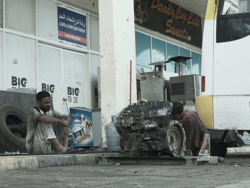 Two men working on a car engine outside a service station, with tools and vehicle parts around them.