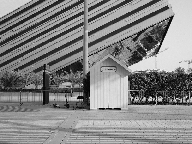 A small white structure stands beside a large, tilted building with colorful stripes in a modern urban setting.