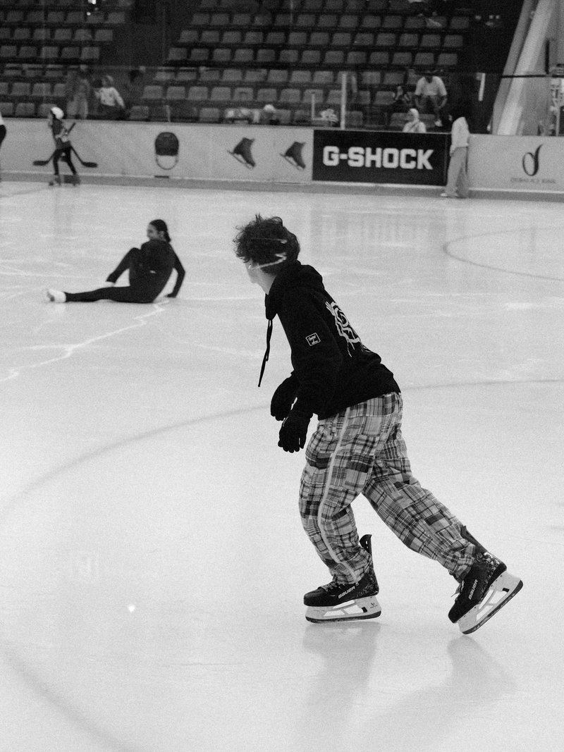 Two ice skaters on an indoor rink; one is skating while the other sits on the ice.