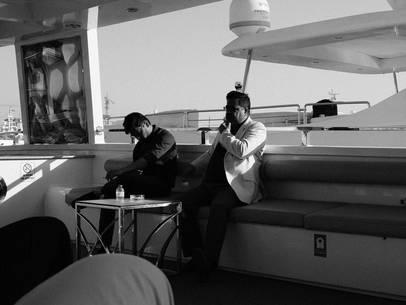 Two men sitting on a boat, one in a black jacket and the other in a white blazer, with a table in between.