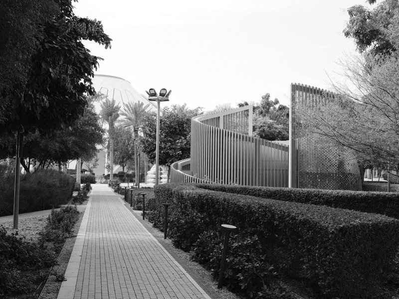 A pathway lined with hedges leading to modern architectural structures under a clear sky.