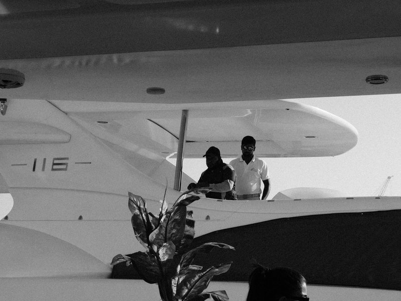 Two people on a boat, with a white yacht in the background and tropical plants in the foreground.