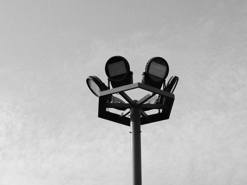 A tall sports light pole with multiple floodlights against a cloudy sky.