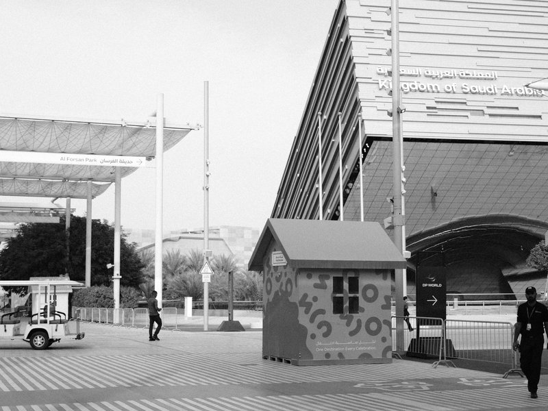 A colorful small house structure near a modern building with a person walking and a vehicle in the scene.