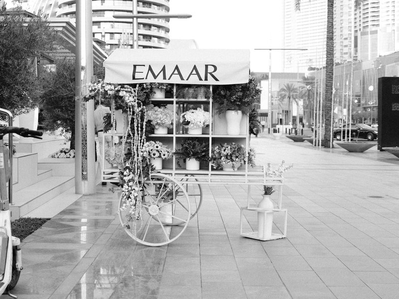 A flower cart with the name 'EMMAR' on a city street, surrounded by modern buildings.