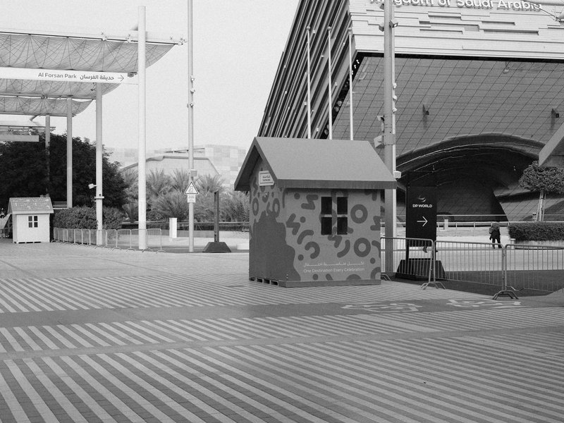 A colorful small house structure in an urban setting with striped pavement and modern architecture in the background.