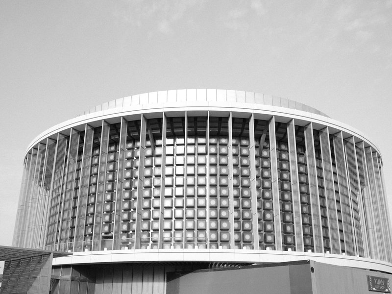A modern circular building with a glass facade and vertical columns against a cloudy sky.