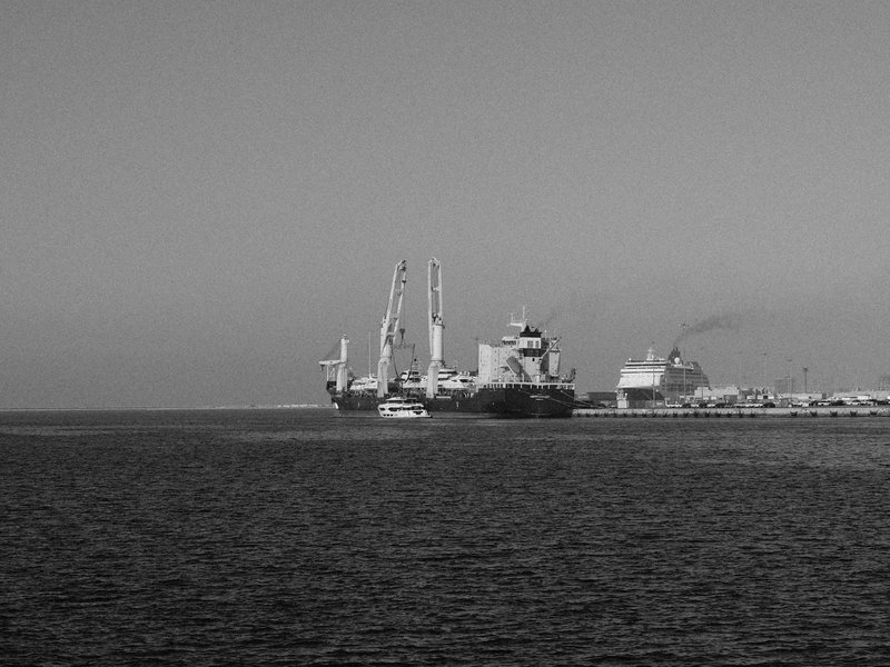 Black and white image of ships docked at a port with cranes and smoke in the background.