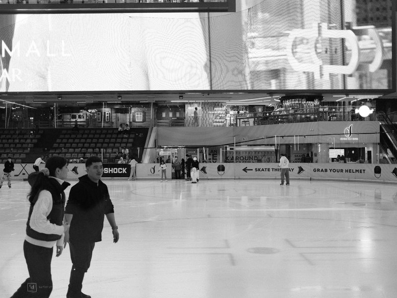 Two skaters on an ice rink with a large screen in the background displaying advertisements.
