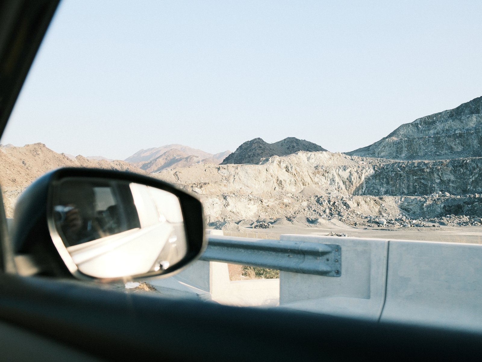 Side mirror of a car reflecting a rocky landscape with mountains in the background under clear skies.