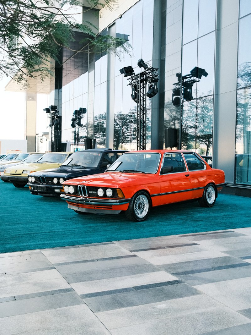 A lineup of vintage cars, including a bright red BMW, in front of a modern glass building.