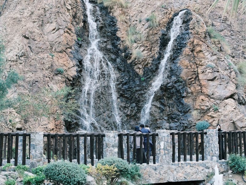 A scenic view of two waterfalls cascading down rocky cliffs, with a stone bridge in the foreground.