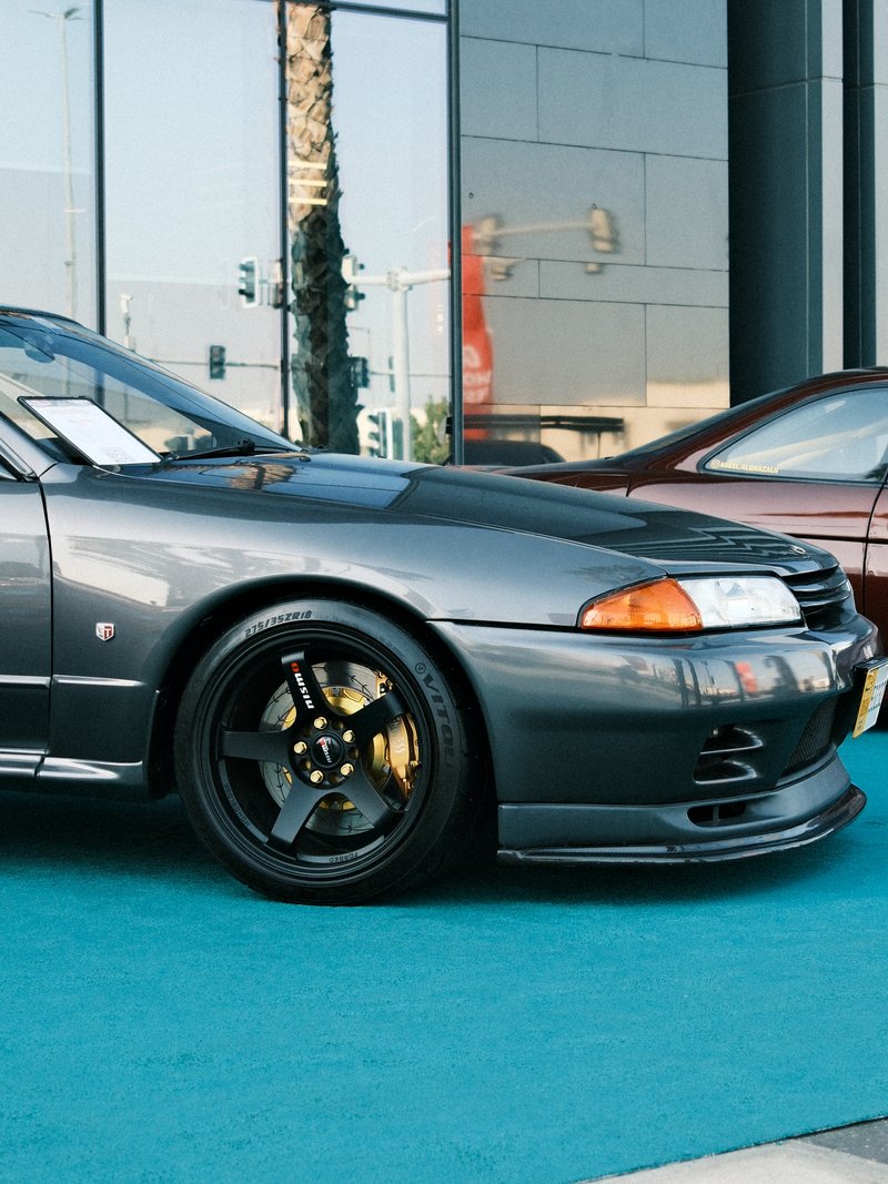 A sleek gray sports car parked on a teal surface with another car in the background.