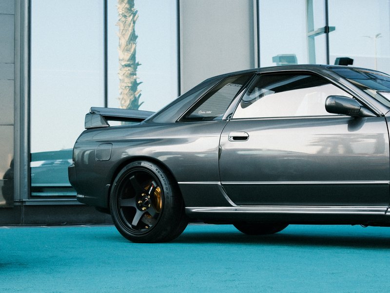 A sleek gray sports car parked beside large glass windows, showcasing its profile and black wheels.