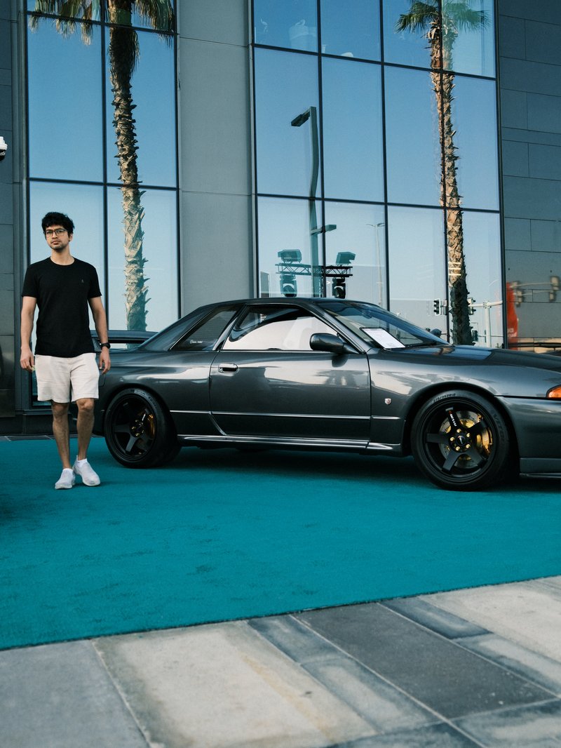 A young man in a black t-shirt and shorts stands beside a sleek gray sports car in front of mirrored buildings.