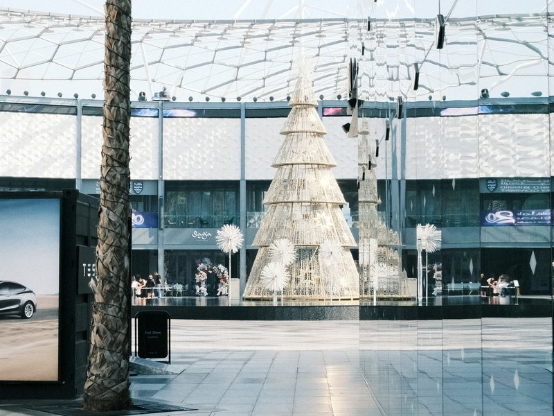 A modern shopping center featuring a large decorative Christmas tree in the center, surrounded by shoppers.