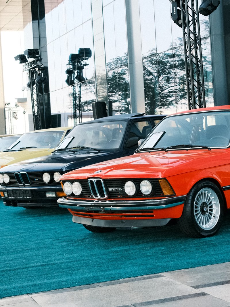 A row of vintage BMW cars in various colors parked on a teal carpet outside a modern building.