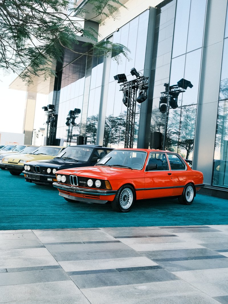 A row of classic cars, including an orange BMW, parked in front of a modern building with large glass windows.