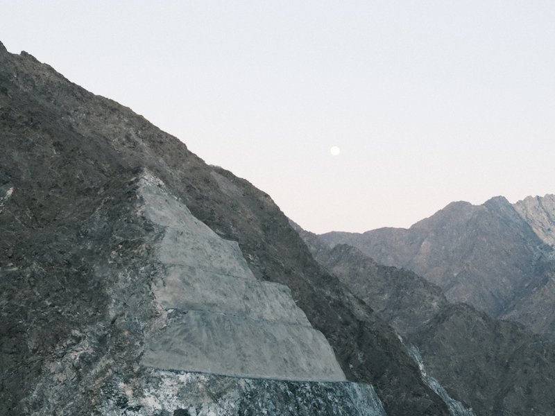 A rocky mountain landscape with a pale moon in the sky, showcasing layered terrain and distant peaks.