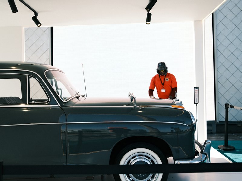A vintage car displayed indoors with a person in an orange shirt standing nearby.