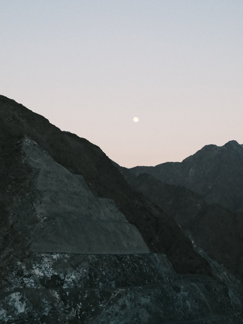 A serene mountain landscape at dusk with a pale moon rising above the peaks.