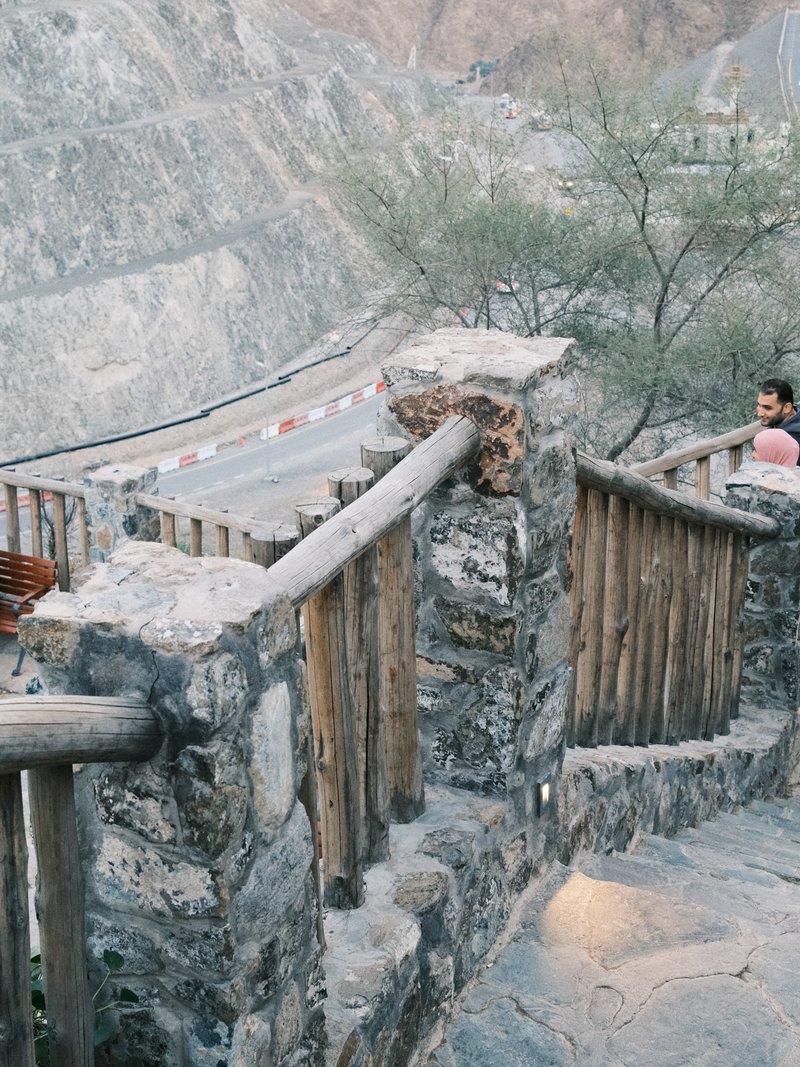 A winding stone staircase with wooden railings leading down a rocky hillside, with a road visible in the background.