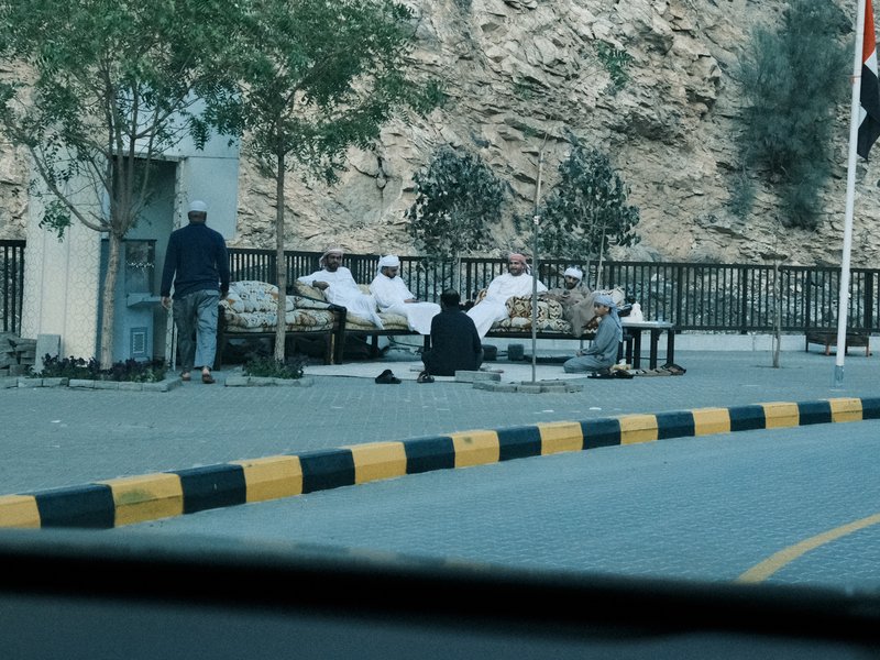 Group of men in traditional attire sitting outdoors near a rocky landscape with trees.