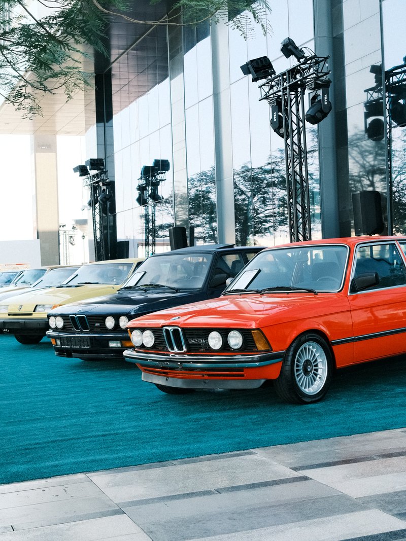A row of vintage cars in various colors parked on a turquoise carpet outside a modern building.
