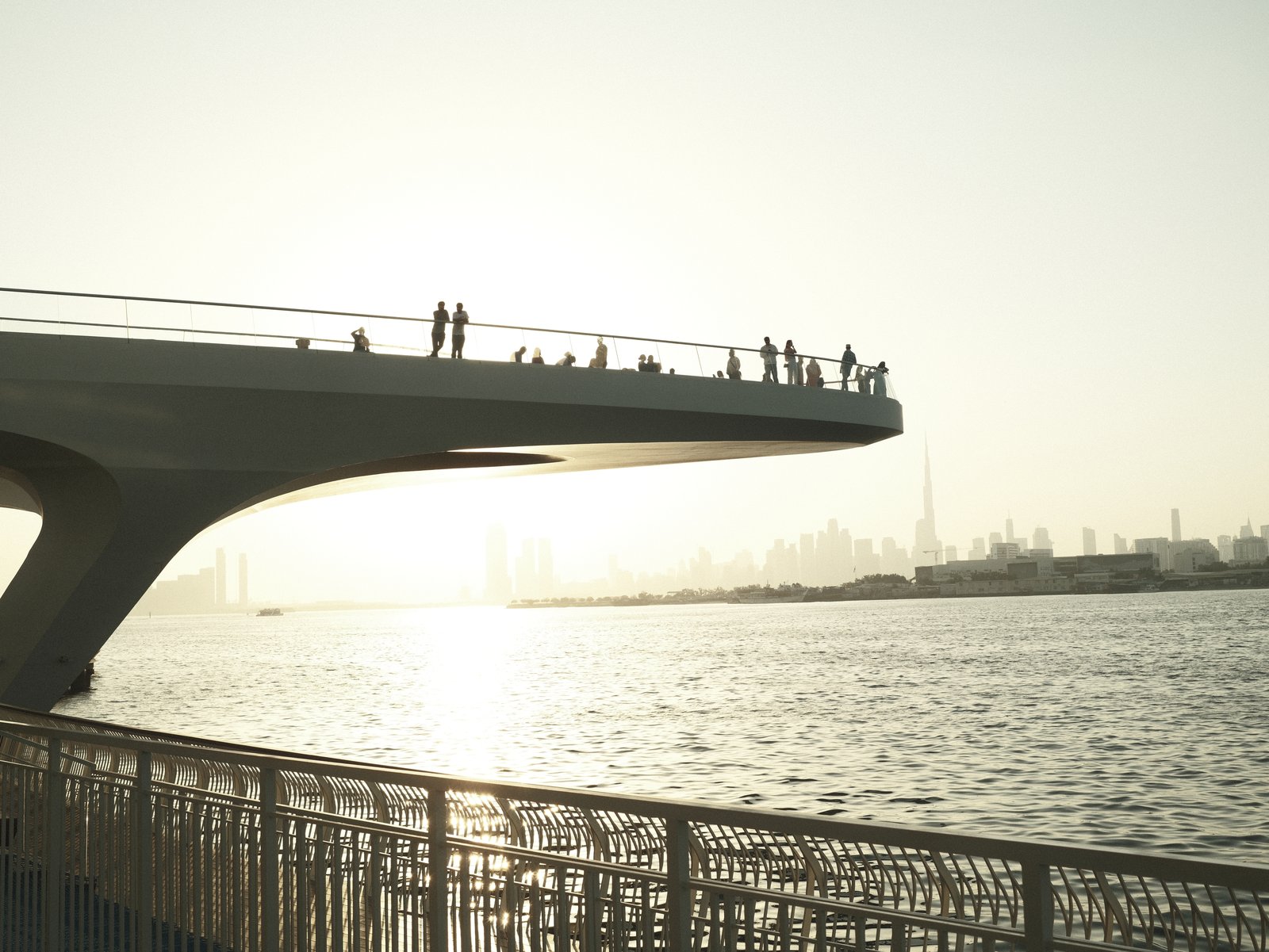 Silhouetted figures on a bridge overlooking a river at sunset, with city skyline in the background.