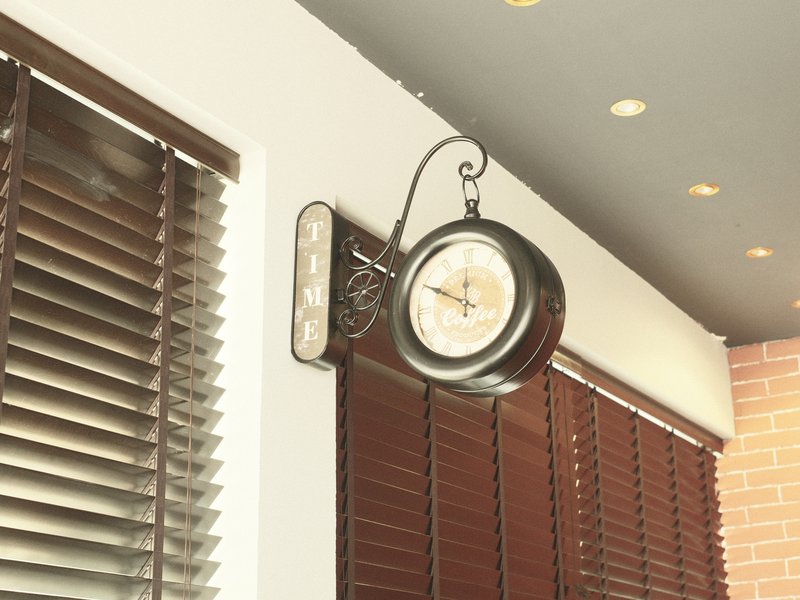 A vintage wall clock hanging in a cafe with wooden blinds and soft lighting.
