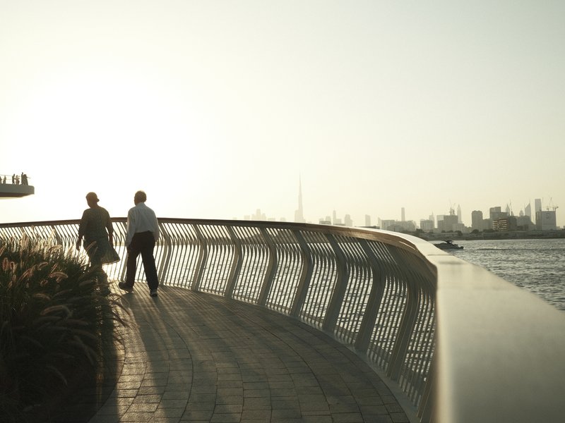 Two people walking along a curved bridge by the water, with a city skyline in the background.