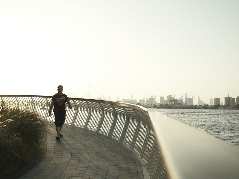A person walking along a curved pathway by the water with a city skyline in the background.