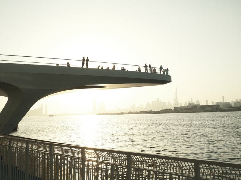 Silhouetted figures on a bridge overlooking a river at sunset, with city skyline in the background.