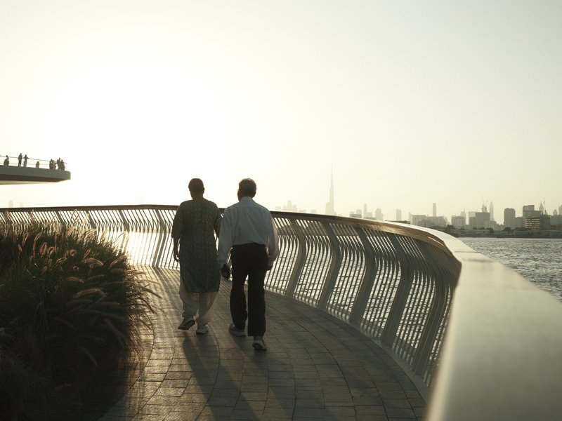 Two people walking along a curved pathway by the water, with a city skyline in the background.