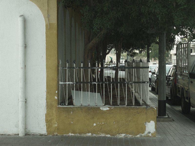 A weathered fence beside a sidewalk, with trees and parked cars in the background.