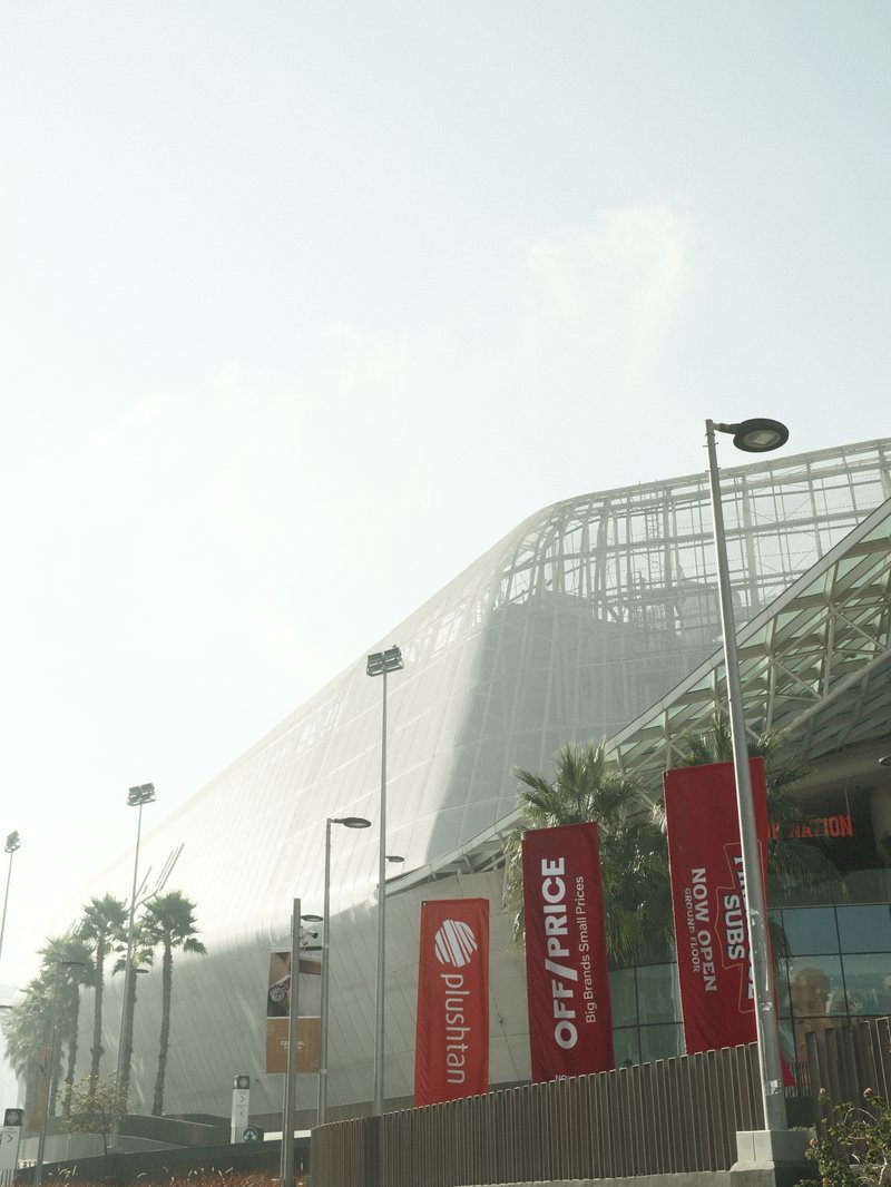 A modern architectural building with a glass facade, palm trees, and banners in a bright, sunny atmosphere.