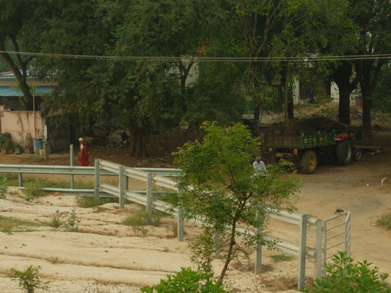 A rural scene featuring a dirt road, trees, and a tractor in the background.