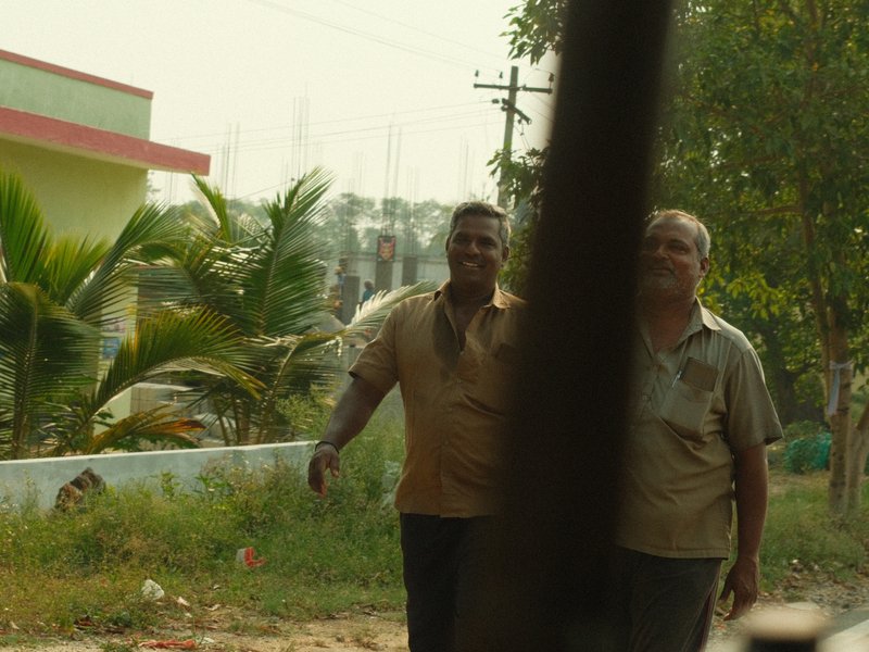 Two men walking along a rural road with greenery and buildings in the background.
