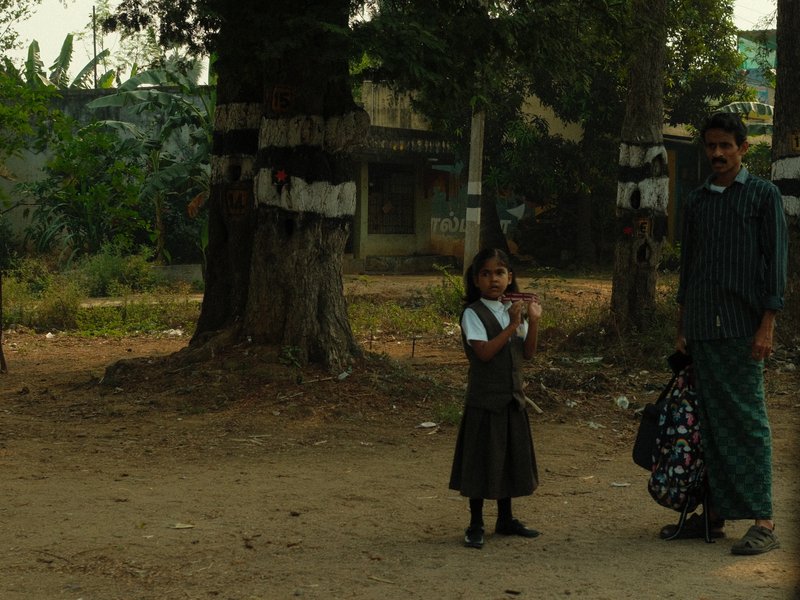 A girl in a school uniform stands with a man in a rural setting, surrounded by trees and a dirt path.