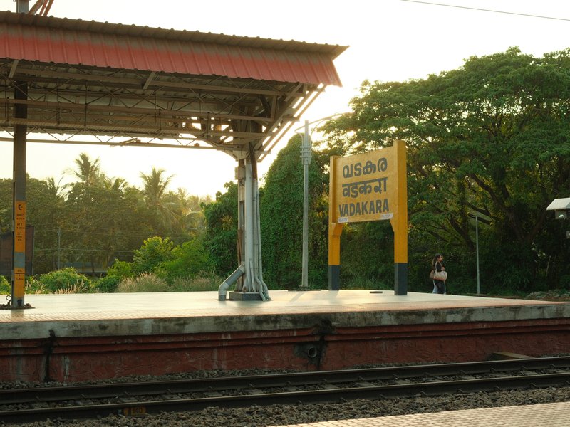 A train station platform with a yellow sign, surrounded by greenery and soft evening light.