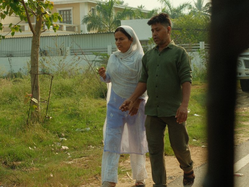 A couple walking hand in hand along a grassy path, surrounded by greenery and buildings in the background.