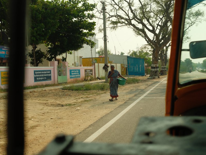 A woman walking along a roadside in a rural area, with trees and buildings in the background.