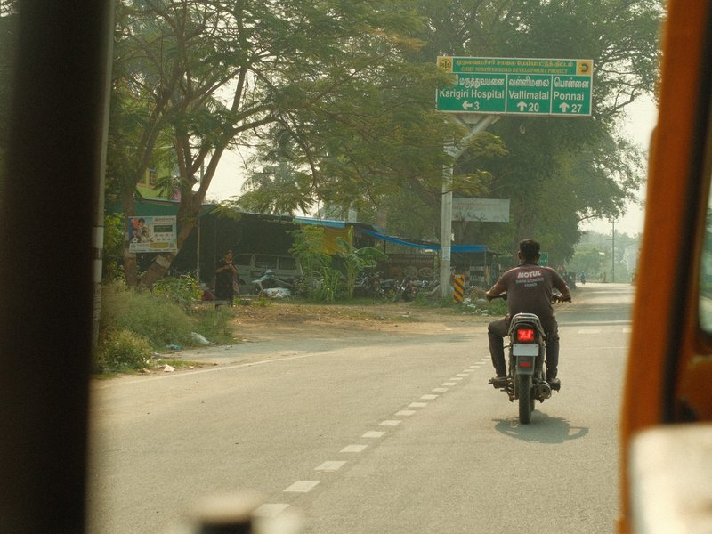 A motorcyclist riding on a rural road with greenery and signage in the background.