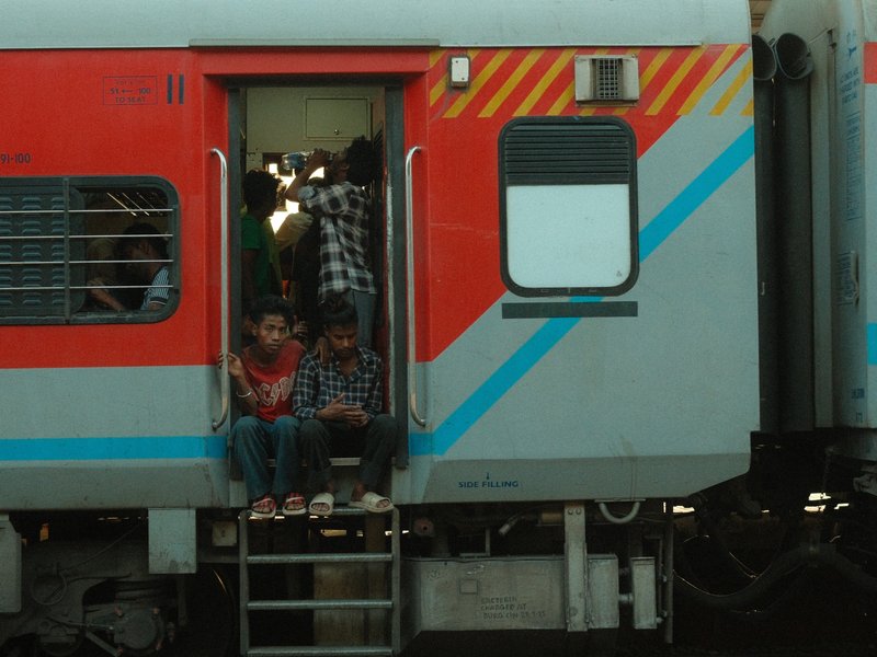 Two men sitting on the steps of a train with others visible inside, urban setting.
