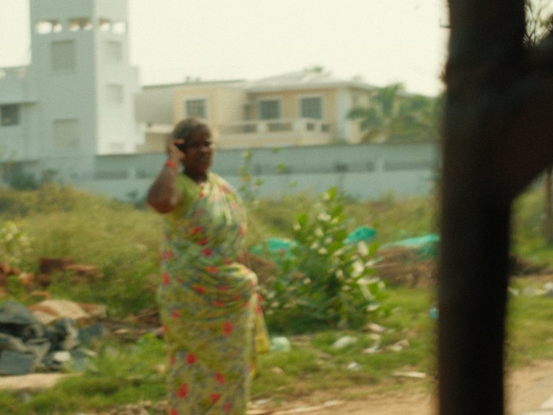 A woman in a colorful sari stands in a grassy area with buildings in the background.