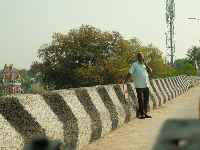A man standing on a patterned concrete barrier with trees and a tower in the background.