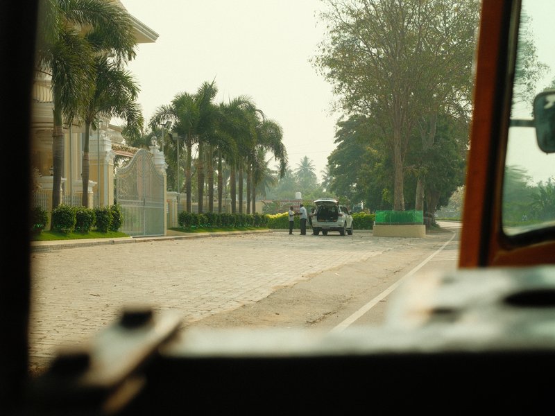 View from a vehicle showing a street lined with palm trees and a vintage car in the distance.