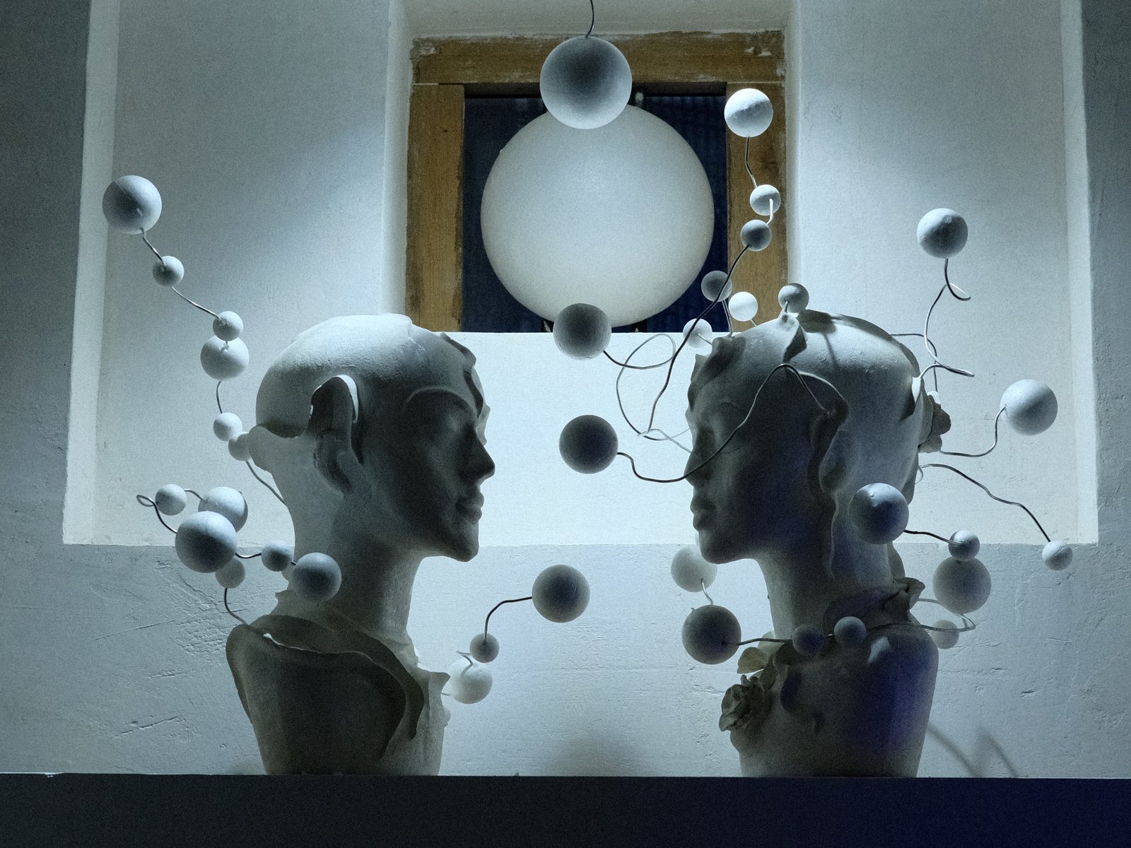 Two white sculptures of heads facing each other with floating spheres around them in a dimly lit space.